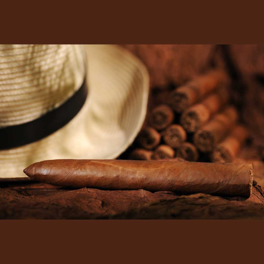 Un sigaro cubano sopra foglie di tabacco, con cappello cubano e altri sigari sullo sfondo