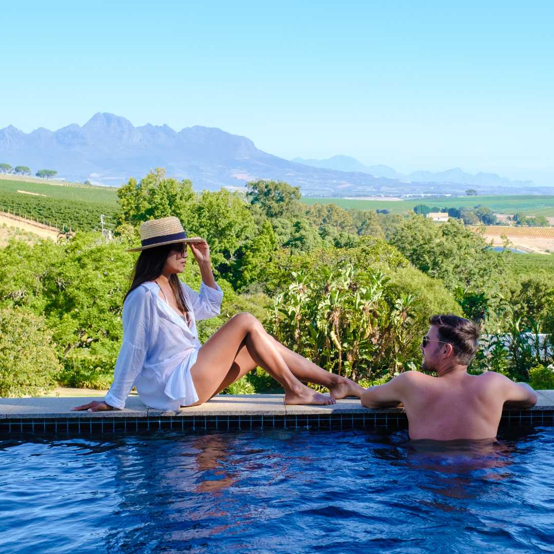 Ein Paar entspannt sich an einem Swimmingpool mit Blick auf eine Weinberglandschaft bei Sonnenuntergang mit Bergen in Stellenbosch, in der Nähe von Kapstadt, Südafrika