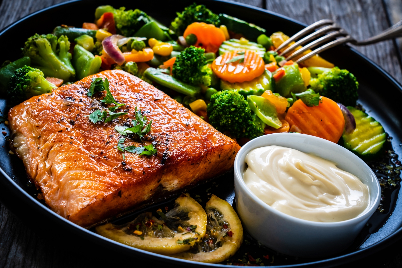 Filete de salmón frito con una mezcla de verduras, limón y mayonesa servido en un plato negro sobre una mesa de madera