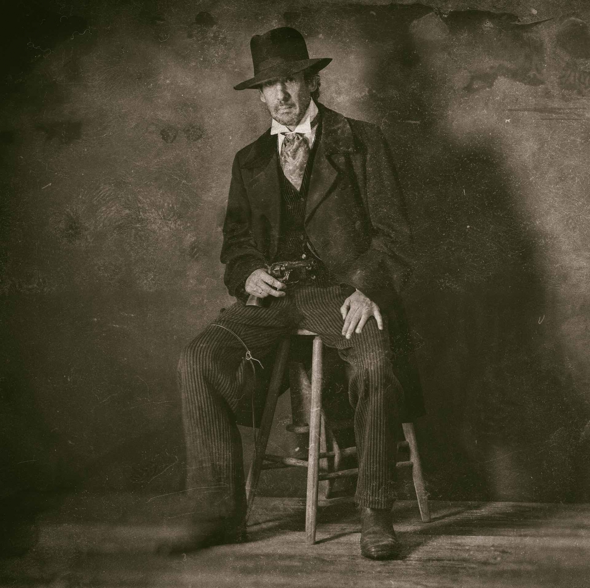 Classica foto di un piatto bagnato di un uomo maturo occidentale vintage del 1900 con rivoltella seduto su uno sgabello di legno.