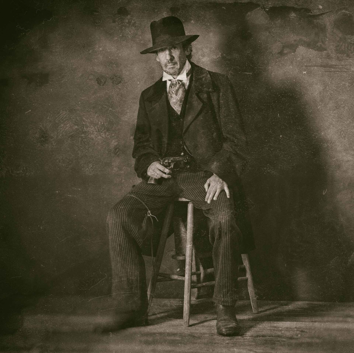 Classic wet plate photo of vintage 1900 western mature man with revolver sitting on wooden stool.