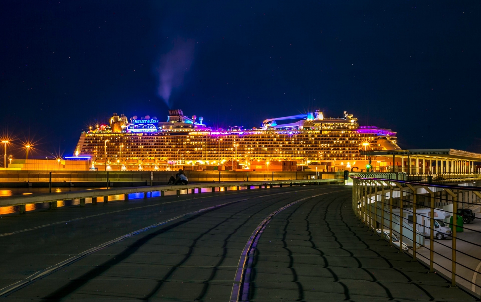 En la vibrante ciudad portuaria de Valencia, un colosal crucero domina el horizonte nocturno y su fachada iluminada se refleja en las brillantes aguas.