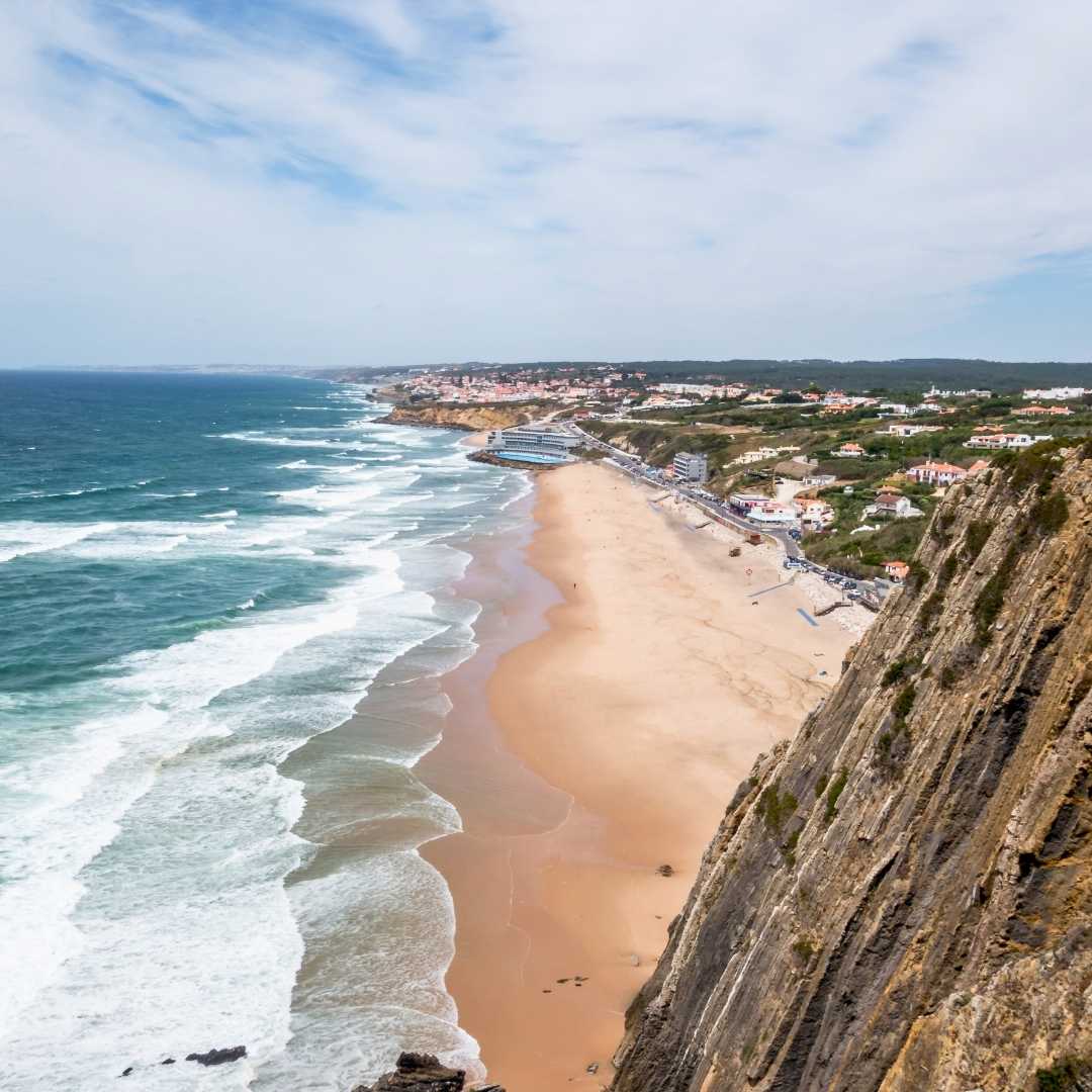 Vista desde los acantilados sobre la playa de arena Praia Grande, cerca de Sintra Portugal en verano con olas rodando bajo un cielo nublado.